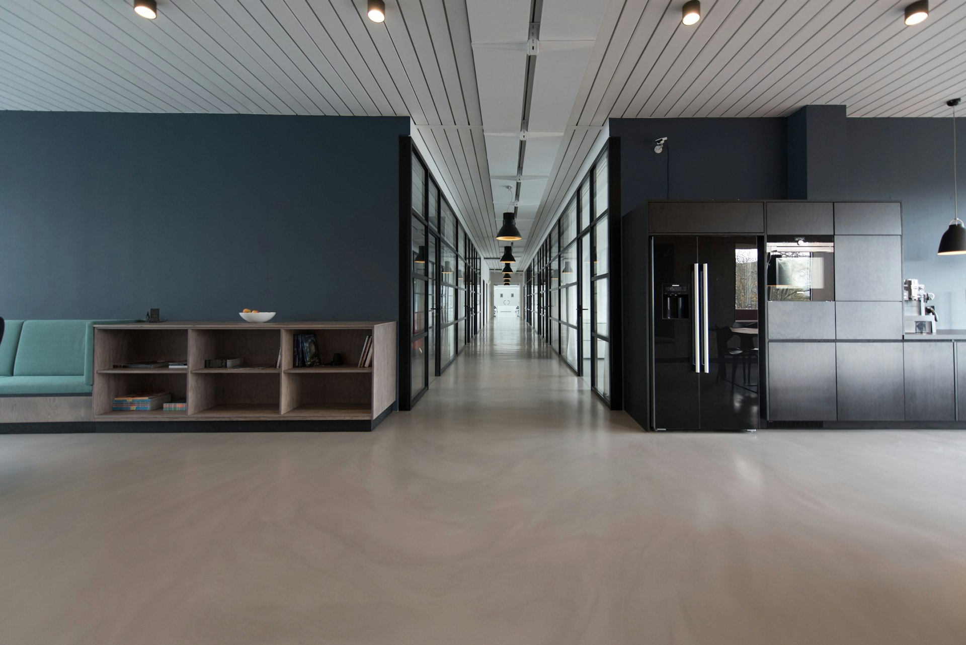 Modern glass-walled office corridor with track lighting and a breakroom kitchen